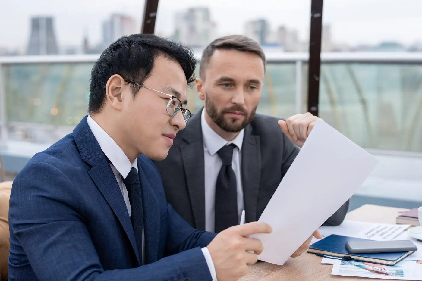 Twee zakenpartners, een Chinese en een Europese man, lezen samen een contract aan een tafel met uitzicht op de stad